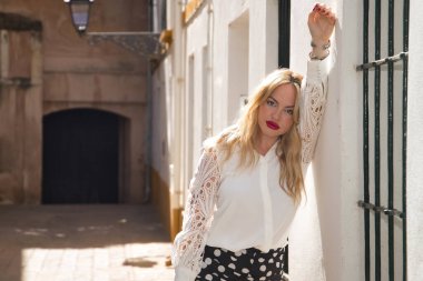 Genç, güzel, sarışın bir kadın, beyaz gömlekli, tatlı ve yumuşak bakışlı, koluna yaslanmış, beyaz bir duvara yaslanmış, güneş ışınlarını alan. Güzelliği, modayı, kadınlığı, eğilimi kavra.