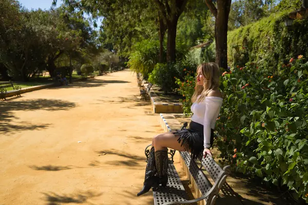 Beyaz tişörtlü, fırfırlı etekli ve kovboy çizmeli, rahat ve sakin, açık hava parkındaki bir bankta oturan genç, güzel, sarışın bir kadın. Güzelliği, modayı, kadınlığı, eğilimi kavra.