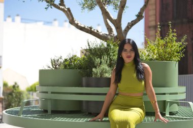 Genç, güzel, esmer, yeşil takım elbiseli Latin bir kadın, rahat ve mutlu yeşil metal bir bankta bitkilerle oturuyor. Güzelliği, modayı, eğilimi, şehri kavra.