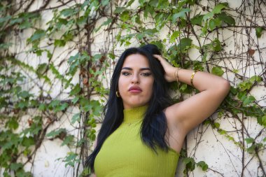 Portrait of young, beautiful, brunette, Latina woman, wearing a green suit, with tender and delicate look, leaning against a wall with a background of ivy. Concept beauty, fashion, trend, city. clipart