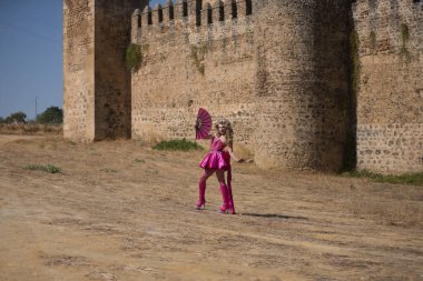 Sürüklenen kraliçe, sarışın, çekici, tacıyla, elbisesiyle, yüksek topuklu botlarıyla ve pembe yelpazesiyle arka planda bir şatoyla dışarı çıkıyor. Gururu, kimliği, çeşitliliği, sanatçıyı kavra.