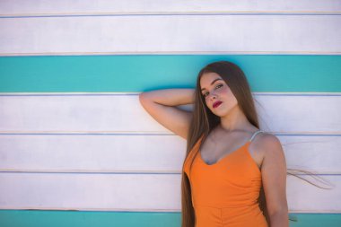 Güzel bir genç kadın portresi, uzun kahverengi saçlı, turuncu bir elbise giyiyor, arkasında bir sahil kulübesi olan tatlı ve şefkatli bir bakışla poz veriyor. Güzelliği, modayı, modayı, kadınlığı kavra.