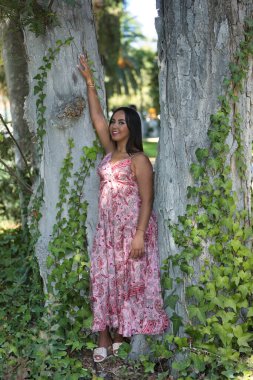 Faslı bir kadın, genç, güzel, koyu renk saçlı, kaşmir bir elbise giymiş, gülümseyen ve mutlu, sarmaşık sarmaşıklı bir ağacın gövdesine yaslanmış. Etnik köken, çeşitlilik, kimlik, güzellik kavramları. Dikey konum.