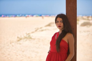Genç, güzel, esmer, uzun saçlı, kırmızı elbiseli, ciddi bakışlı, arka planda plajı olan tahta bir direğe yaslanan bir kadın. Güzelliği, modayı, modayı, kadınlığı kavra.