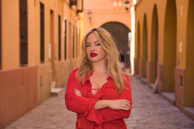 Genç, güzel, sarışın, kırmızı elbiseli, kollarını kavuşturmuş, kameraya bakan, özgür ve güçlü bir kadın portresi. Kavram güçlendirme, başarı, kadınlık, özgürlük.