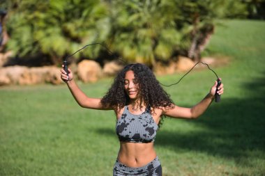 Latin bir kadın, genç, güzel, esmer, kıvırcık saçlı ve spor elbiseli, komik bir tavırla ip atlarken saçlarını hareket ettiriyor. Konsept spor, egzersiz, atlama, aktif.