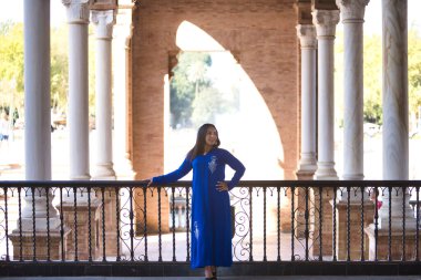 Faslı bir kadın, genç, güzel, esmer, mavi abaya 'lı, dikkati dağınık bakışlı, sütunlar arasındaki korkuluklara yaslanmış. Etnik köken, çeşitlilik, kadınlık, güzellik kavramları.