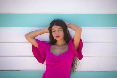 Genç, güzel, esmer, uzun saçlı, pembe takım elbiseli, elleri başının arkasında, arkasında bir plaj kulübesi olan kameraya bakan bir kadın. Güzelliği, modayı, kadınlığı kavra.