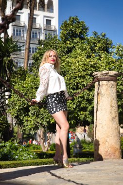 Genç, güzel, sarışın bir kadın, beyaz gömlekli ve siyah benekli etekli ve altın topuklu, kameraya bakıyor, iki kolon arasında büyük metal bir zincir üzerinde oturuyor. Dikey konum.