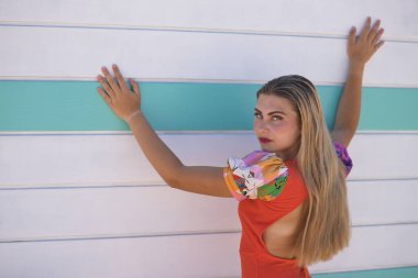 Genç bir kadın, güzel, mavi gözlü, sarışın, turuncu renkli elbiseli, arkası dönük, omzunun üzerinden bakıyor, arka planda bir sahil kulübesine yaslanıyor. Güzelliği, modayı, eğilimi, kadınlığı kavra.