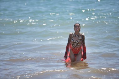 Genç bir kadın, güzel, esmer, leopar mayosu ve elbisesi var, sırılsıklam, arka planda deniz ile sahilin kıyısında yürüyor. Güzellik kavramı, kadınlık.