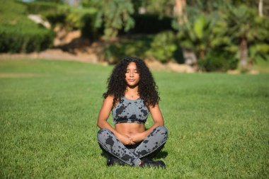 Genç, güzel, esmer, kıvırcık saçlı ve spor giyimli Latin bir kadın, açık hava parkında yoga egzersizleri yapıyor. Konsept spor, rahatlama, meditasyon, egzersiz.