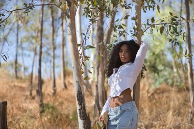 Latin bir kadın, genç, güzel, siyah saçlı, kıvırcık saçlı, beyaz gömlekli ve kot pantolonlu, bir okaliptüsün gövdesine yaslanmış, kameraya bakıyor, rahat ve sakin. Barışı kabul et, rahatla..