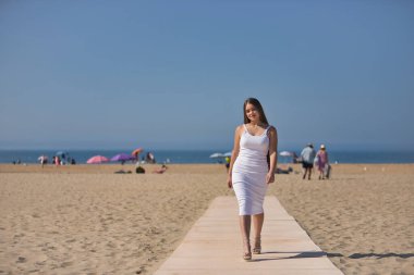 Beyaz elbiseli ve topuklu ayakkabılı genç güzel bir kadın sahilde tahta bir geçitte geçit töreni yapıyor. Güzelliği, modayı, modayı, kadınlığı kavra.