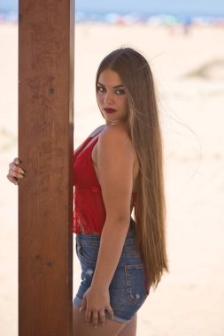 Güzel bir genç kadının portresi, uzun kahverengi saçlı, kırmızı bluzlu ve kot pantolonlu, kameraya bakarak poz veren, arka planda plajı olan tahta bir direğe tutunan. Güzellik kavramı, kadınlık.