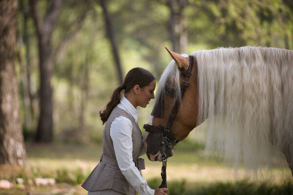 Молодая женщина, одетая как всадница, складывает лоб вместе со своей красивой коричневой лошадью с белой гривой посреди леса. Концепция животных, верховая езда, конный спорт, лошадь.