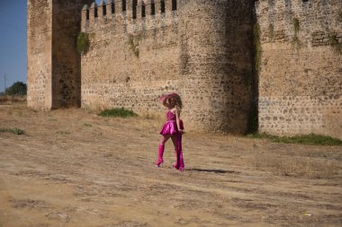 Sürükleme kraliçesi, sarışın, çekici, tacıyla, elbisesiyle, yüksek topuklu botlarıyla ve pembe yelpazesiyle, arka planda kalesi olan açık havada yelpaze ile kendini güneşten koruyor. Gururu kavra.