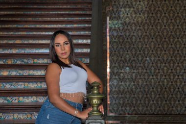 Faslı bir kadının portresi, genç, güzel, esmer, beyaz bluzlu, kot pantolonlu, kameraya bakan, Sevilla Meydanı 'ndaki güzel bir merdivenin korkuluğuna dayanan.