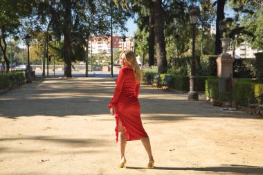 Genç, güzel, sarışın, kırmızı elbiseli, altın topuklu, ayakta, gülümseyen ve mutlu, açık hava parkının ortasında sırt üstü poz veren bir kadın. Güzellik kavramı, moda, kadınlık, moda.