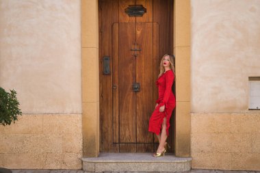 Genç, güzel, sarışın, kırmızı elbiseli, altın topuklu, kameraya bakıyor, güzel bir kapı çerçevesine yaslanıyor. Güzellik kavramı, moda, kadınlık, moda.