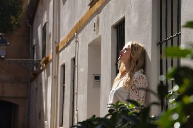 Beyaz gömlekli, genç, güzel, sarışın bir kadın, beyaz bir duvara yaslanmış, güneş ışınlarını alan gökyüzüne bakıyor. Güzelliği, modayı, kadınlığı, eğilimi kavra.