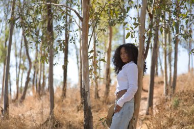 Latin bir kadın, genç, güzel, siyah saçlı, kıvırcık saçlı, beyaz gömlekli ve kot pantolonlu, bir okaliptüsün gövdesine yaslanmış, kameraya bakıyor, rahat ve sakin. Barışı kabul et, rahatla..