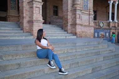 Faslı bir kadın, genç, güzel, esmer, beyaz bluzlu ve kot pantolonlu, rahat ve sakin bir şekilde sonsuzluğa bakıyor, bazı basamaklarda oturuyor. Etnik köken kavramı, çeşitlilik, kadınlık, güzellik.