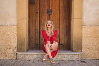 Genç, güzel, sarışın, kırmızı elbiseli ve altın topuklu ayakkabılı bir kadın kameraya bakıyor, güzel bir kapının pencere eşiğinde oturuyor. Güzellik kavramı, moda, kadınlık, moda.