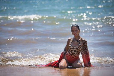 Genç bir kadın, güzel, esmer, leopar mayo ve sarong giymiş, dizlerinin üstünde, kumsalın sahilinde sırılsıklam poz veriyor, arka planda deniz var. Güzelliği, modayı, kadınlığı kavra.