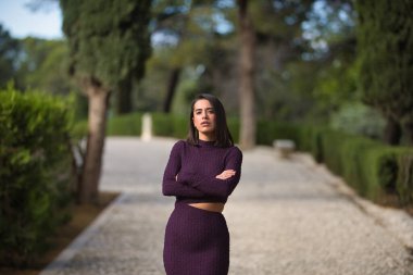 Genç, güzel, esmer, İspanyol bir kadın, mor bir elbise giyiyor, kolları çapraz, güçlü ve bağımsız, selvi ağaçlarıyla çevrili taş bir yolda. Güzelliği, modayı, modayı kavra.