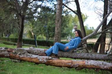 Genç, güzel, esmer, İspanyol bir kadın, kot elbise giyiyor, rahat ve sakin bir şekilde kameraya bakıyor, bir ağacın gövdesinde yatıyor, doğada. Kavram moda, güzellik, moda, kadınlık.