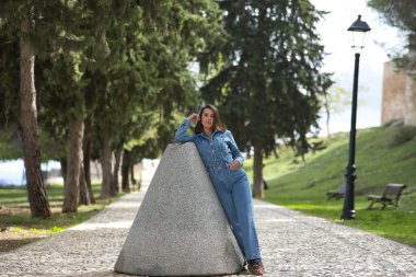 Genç, güzel, esmer, İspanyol bir kadın, kot elbise giyiyor, tatlı ve çekici poz veriyor, taş bir yolda eski bir değirmen taşına yaslanıyor. Kavram moda, güzellik, moda, kadınlık.