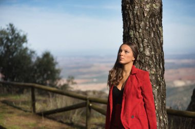 Kırmızı ceketli ve pantolonlu genç ve güzel bir kadın, siyah bluzlu, gökyüzüne bakıyor, Tanrı 'nın doğadaki bir ağacın gövdesine yaslandığını düşünüyor. Güzelliği, modayı, dini, kadınlığı kavra.