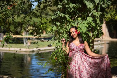 Faslı bir kadın, genç, güzel, esmer, kaşmir elbiseli, kulağında yeşil yapraklarla çevrili bir çiçek, bir gölün yanında. Etnik köken, çeşitlilik, kimlik, güzellik kavramı.