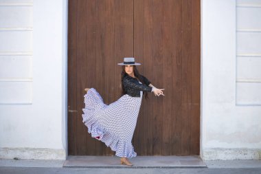 Young woman, beautiful, brunette, flamenco dancer, wearing a beautiful dress with jacket and hat, dancing flamenco with a wooden door in the background. Flamenco concept, typical, Spanish, Seville. clipart