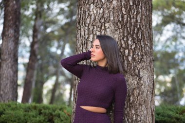 Genç, güzel, koyu saçlı, İspanyol bir kadının portresi, mor bir elbise giymiş, dikkati dağılmış ve hayalperest bakışlı, büyük bir ağacın gövdesine yaslanmış. Güzelliği, modayı, eğilimi kavra.