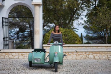 Genç, güzel, esmer, gri elbiseli İspanyol kadın sepetli antika bir motosiklet kullanıyor, dikkati dağınık görünüyor. Konsept motor, motosiklet, vintage, moda, güzellik.