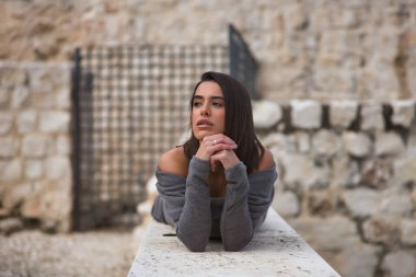 Genç, güzel, esmer, gri elbiseli İspanyol bir kadının portresi, üzgün ve dikkati dağınık, elleri çenesinde, taş bir duvarda yatıyor. Kavram moda, güzellik, moda, kadınlık.