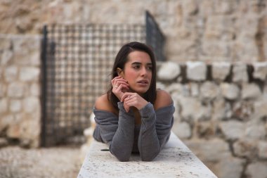 Genç, güzel, esmer, gri elbiseli İspanyol bir kadının portresi, üzgün ve dikkati dağınık, elleri çenesinde, taş bir duvarda yatıyor. Kavram moda, güzellik, moda, kadınlık.