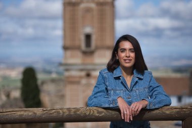 Genç, güzel, esmer, İspanyol kadın portresi, kot pantolon giymiş, özgür ve güçlü, arka planda şehir manzarası olan ahşap korkuluklara yaslanmış. Kavram trendi, kadınlık.