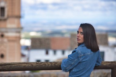 Genç, güzel, esmer, İspanyol kadın portresi, kot giyiyordu, utangaç ve çekingendi, arkası dönük, ahşap bir korkuluğa yaslanmıştı, arka planda kentsel bir manzara vardı..