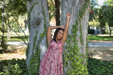 Faslı bir kadın, genç, güzel, koyu renk saçlı, kaşmir bir elbise giymiş, gülümseyen ve mutlu bir şekilde kameraya bakıyor, sarmaşık asmalarıyla bir ağacın gövdesine yaslanıyor. Etnik köken, çeşitlilik, kimlik kavramı.