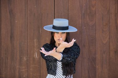 Portrait of young woman, beautiful, brunette, flamenco dancer, wearing a beautiful dress with jacket and hat, doing flamenco poses with hands, with a wooden door in the background. clipart