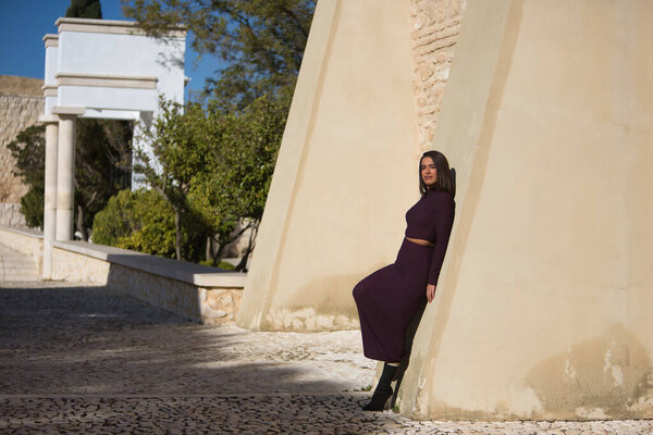 Young, beautiful, brunette, Hispanic woman, wearing a purple dress and boots, leaning against a wall, looking at the camera, receiving the sun's rays. Concept beauty, fashion, trendy, loneliness.