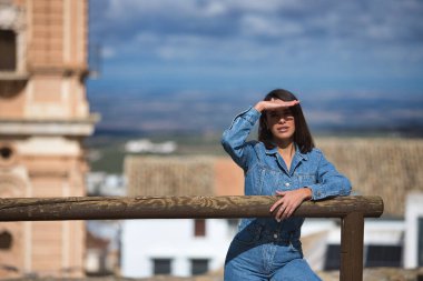 Genç, güzel, esmer, İspanyol bir kadın, kot elbise giyiyor, güneş ışınlarından gözlerini eliyle kapatıyor, arka planda şehir manzarası olan ahşap bir korkuluğa yaslanıyor..