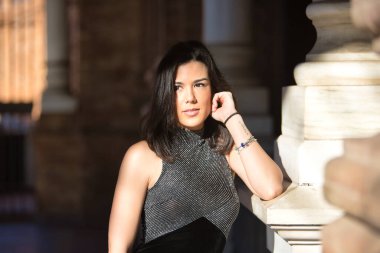 Genç, güzel, esmer kadın portresi, siyah bir fantezi elbisesi, rüya gibi bir görünüşü var, mermer bir sütuna yaslanmış, güneş ışınlarını alıyor. Güzelliği, modayı, modayı, kadınlığı kavra.