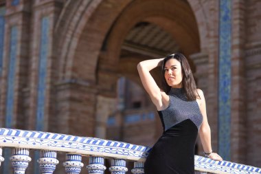 Süslü siyah elbiseli, genç, güzel, esmer kadın, sonsuzluğa bakıyor, Seville 'de İspanya meydanındaki bir köprüde fayanslı bir korkuluğa yaslanıyor. Güzellik, moda, kadınlık, turizm kavramları.