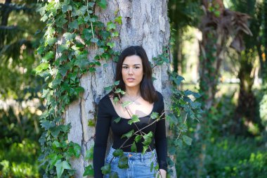 Siyah tişört ve kot pantolon giymiş, bir ağacın gövdesine yaslanmış ve doğayla bağlantılı, sarmaşığa dolanmış genç, güzel, koyu renk saçlı bir kadın. Güzellik kavramı, kadınlık, doğa, bitkiler, ağaçlar.