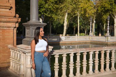 Faslı bir kadın, genç, güzel, esmer, beyaz bluzlu ve kot pantolonlu, sonsuzluğa bakıyor, bir taş korkuluğa yaslanıyor. Etnik köken kavramı, çeşitlilik, kadınlık, güzellik.