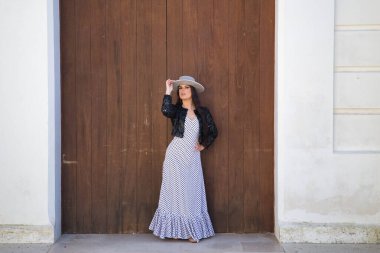 Genç bir kadın, güzel, esmer, flamenko dansçısı, ceketli ve şapkalı güzel bir elbise giyiyor, arka planda ahşap kapılı poz veriyor. Flamenko konsepti, dansçı, tipik, İspanyolca, Sevilla.